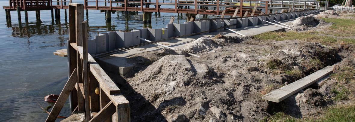 Seawall breakwall construction strengthening work on large body of water - stock photo  Seawall break wall concrete strengthening work construction site utilizing wood framing on large body of water with docks, boats and houses in the background on sunny.